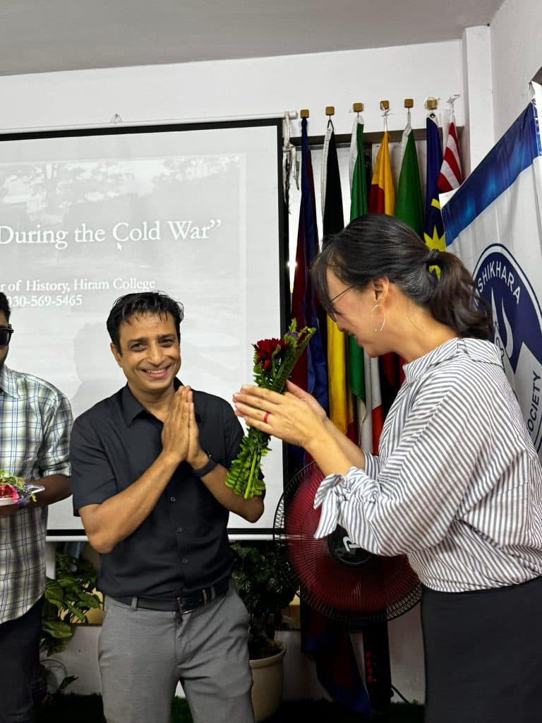 A speaker greeted with flowers in front of a lecture screen and a row of international flags