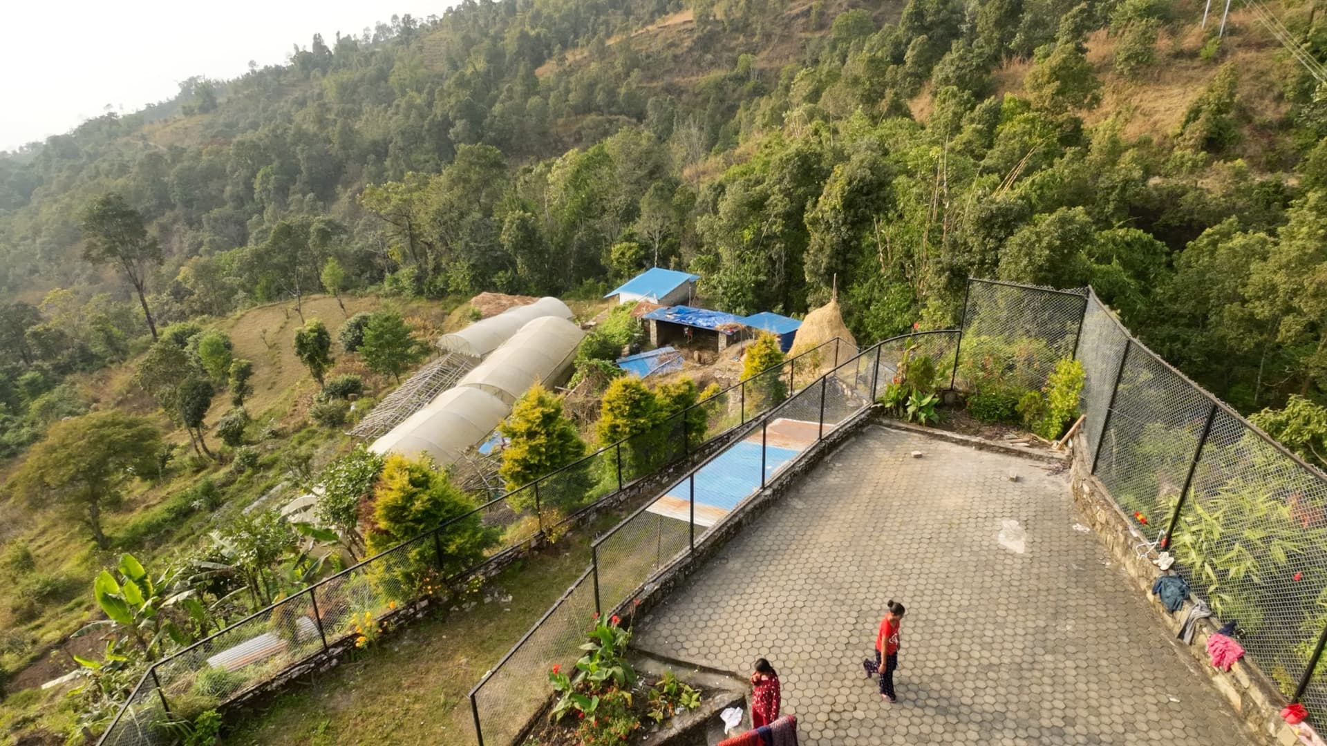Aerial view of a fenced hillside community site with polytunnels, a paved courtyard and a covered structure