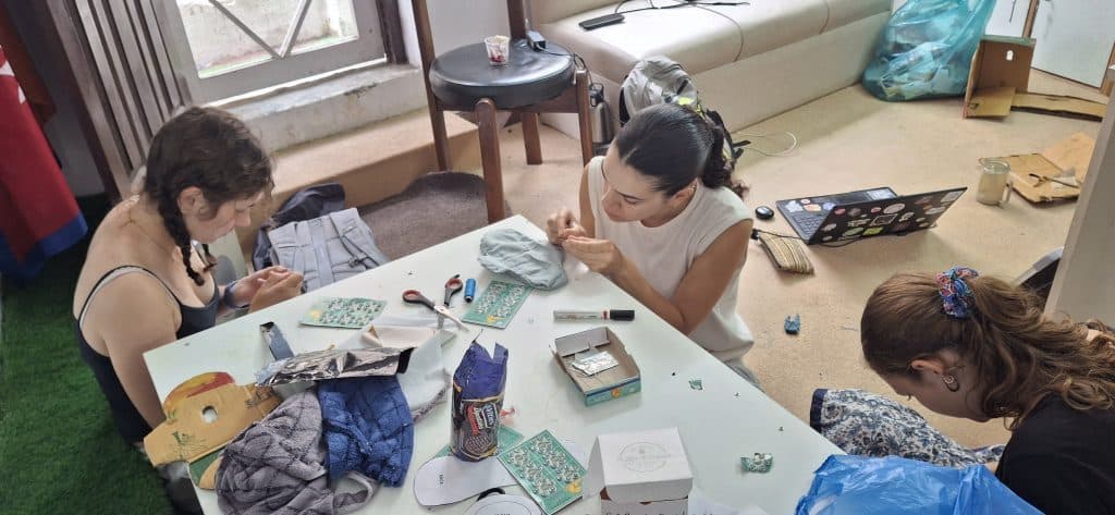 Three people working with fabric, thread and small tools spread across a glass table