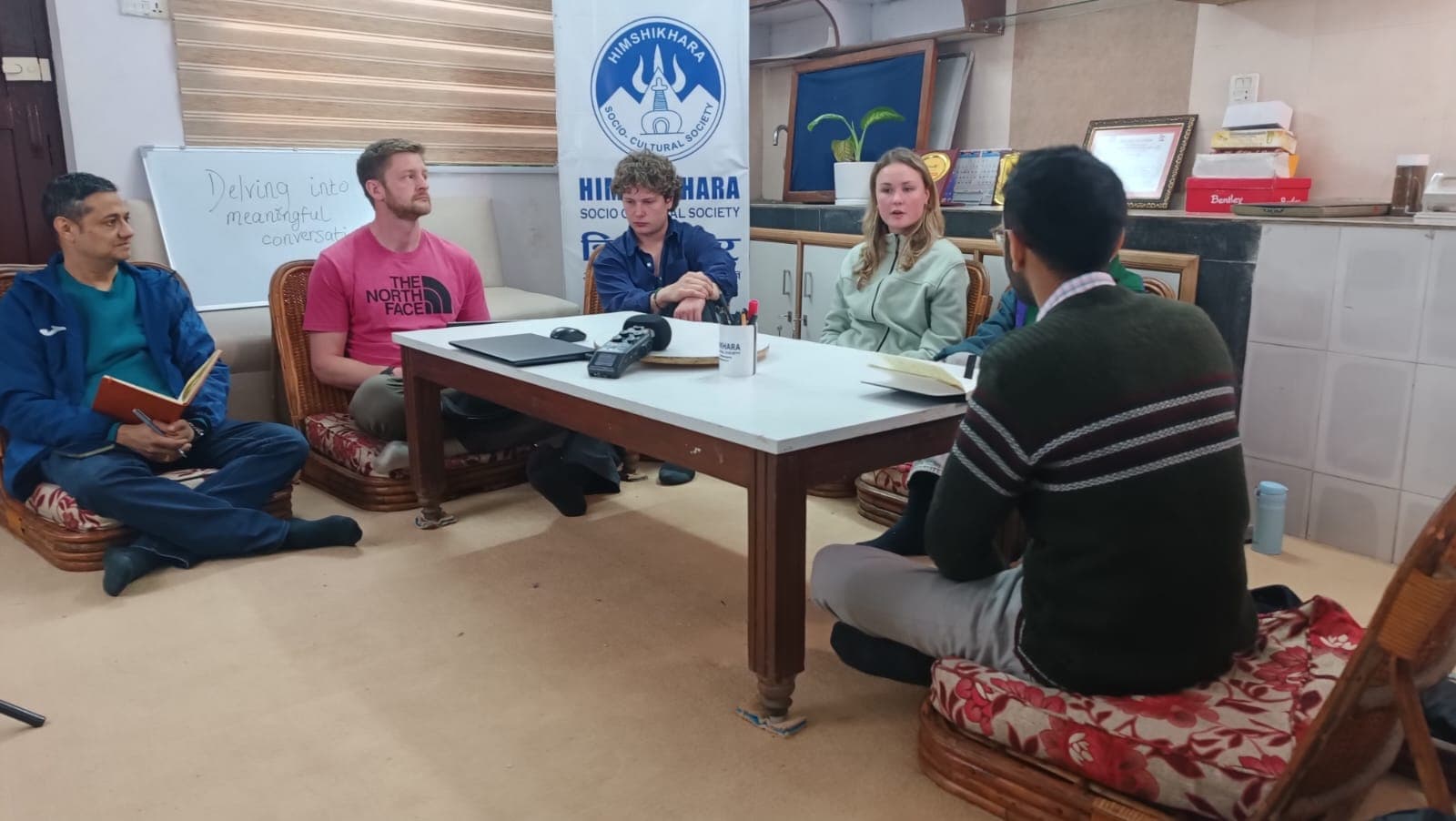 Five people seated around a low table with microphones during a recorded discussion, an organisation banner behind them