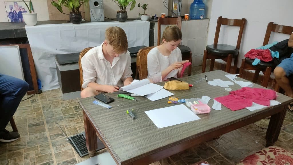 Two volunteers at a wooden table measuring and cutting pink fabric with paper templates