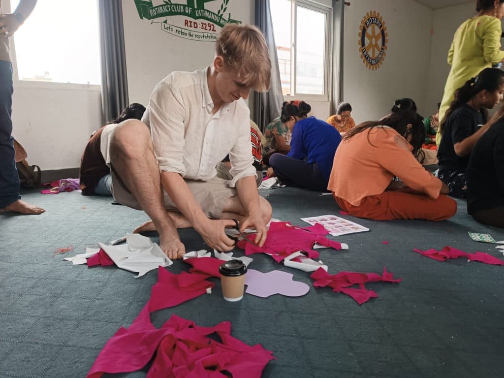 A volunteer cutting pink fabric into reusable pad shapes on the floor of a hall while participants work nearby