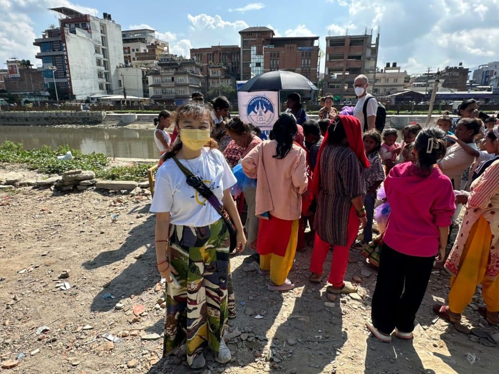 Women and children gathered beside a river under a branded shelter during a community support activity