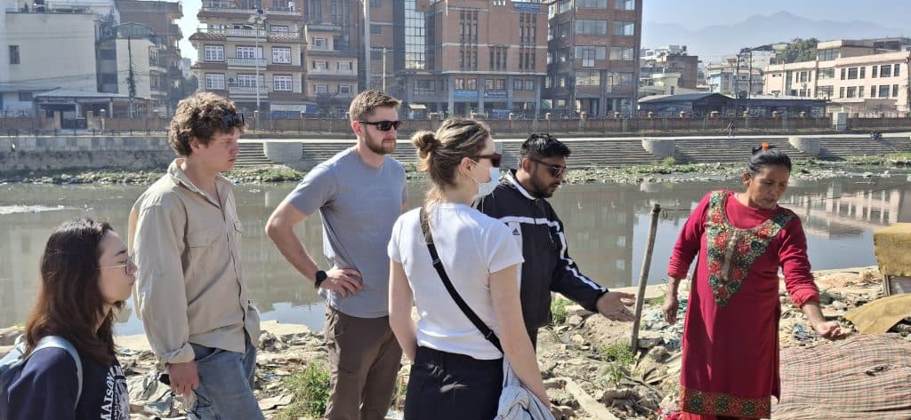 A community member speaking with a small group beside a river with the city behind them