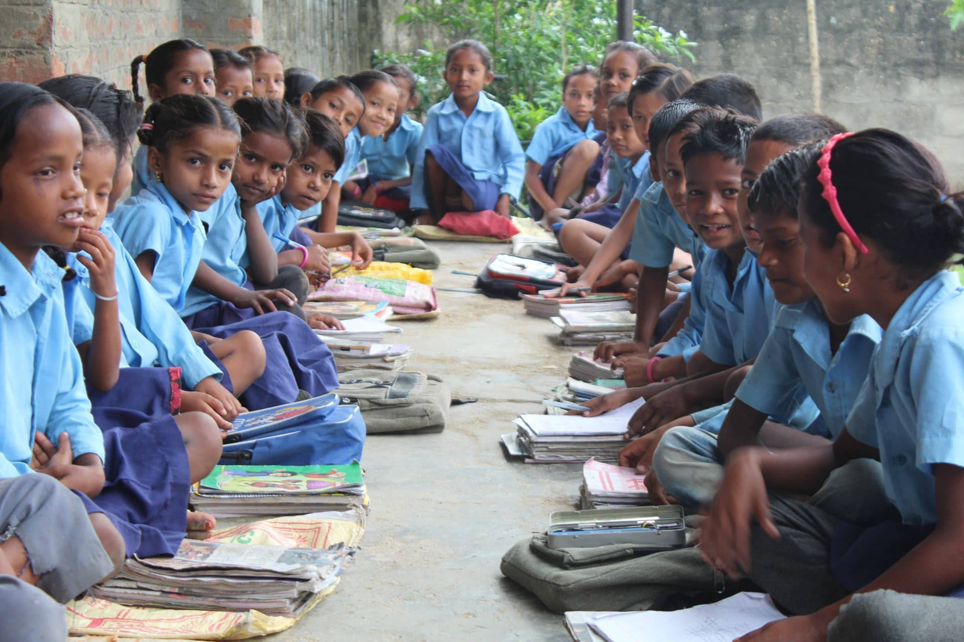 Rows of school students in blue uniforms seated on the ground with open books and school bags