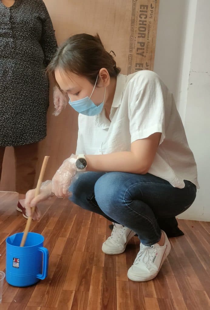 A participant crouching on the floor, stirring soap mixture in a blue container with a wooden stick