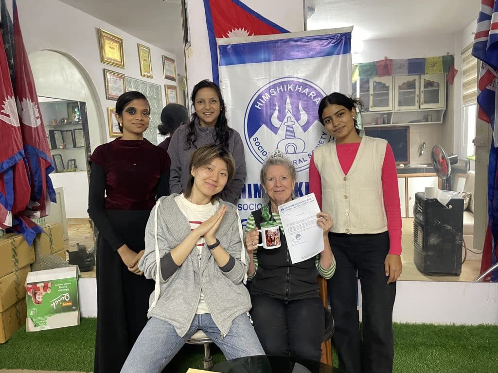 Five volunteers and staff members posing together in an office in front of an organisation banner and Nepali flags