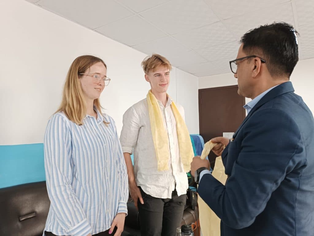 Two visitors being welcomed with a ceremonial scarf by a member of staff in an office